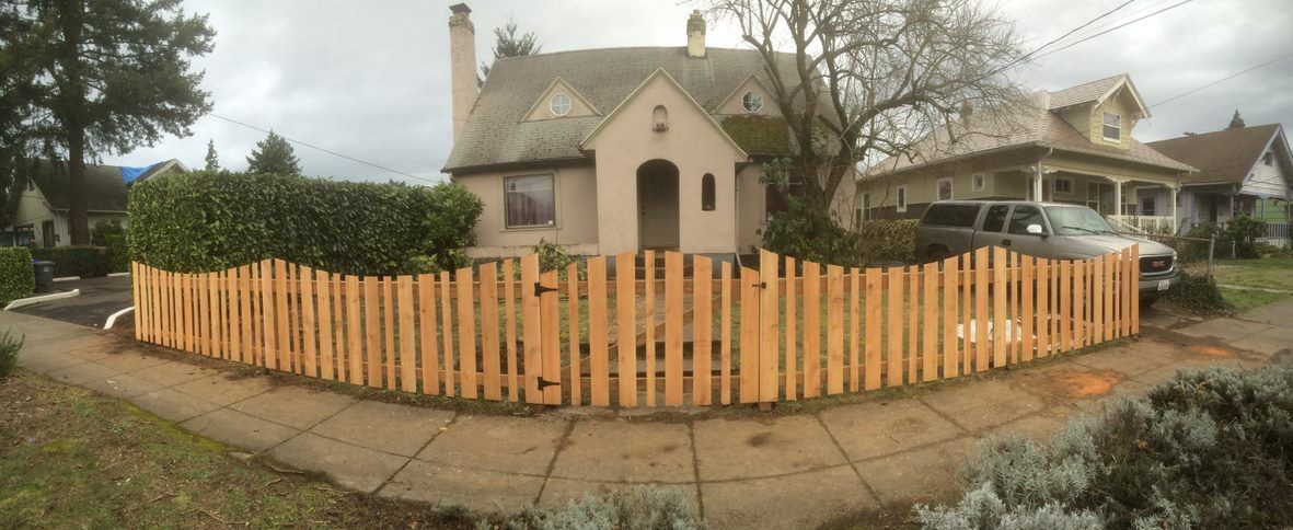 Yellow picket fence surrounds a house and yard in a suburban neighborhood.