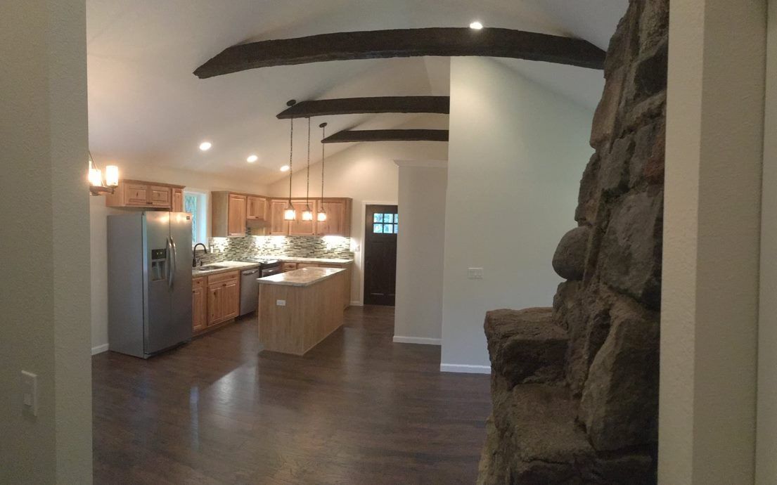 Open-concept kitchen and living area with wood beams, stainless fridge, and stone fireplace.