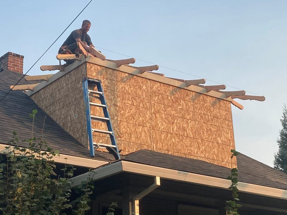 Worker on a roof installing plywood siding beside a blue ladder on a house under construction