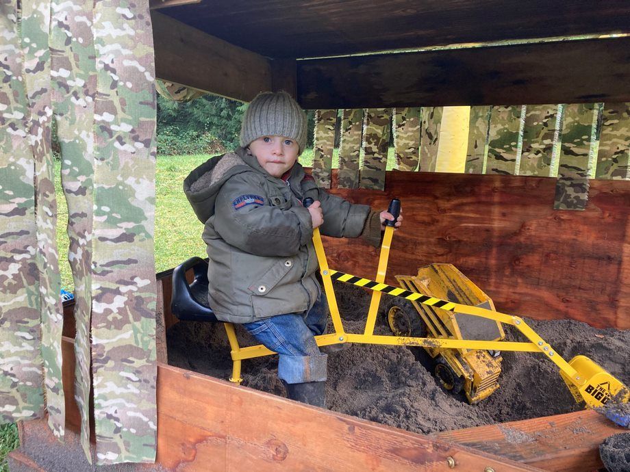 Child in a knit hat sits on a yellow toy tractor inside a wooden play structure.