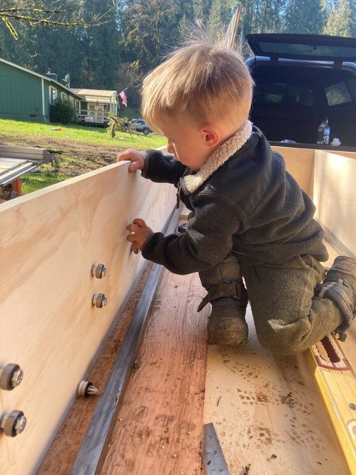 Child kneeling in a trailer, examining a wooden panel with metal bolts outdoors.