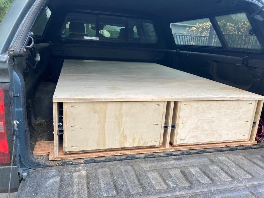 Pickup truck bed with two large wooden storage boxes and a flat plywood top, viewed from the open tailgate