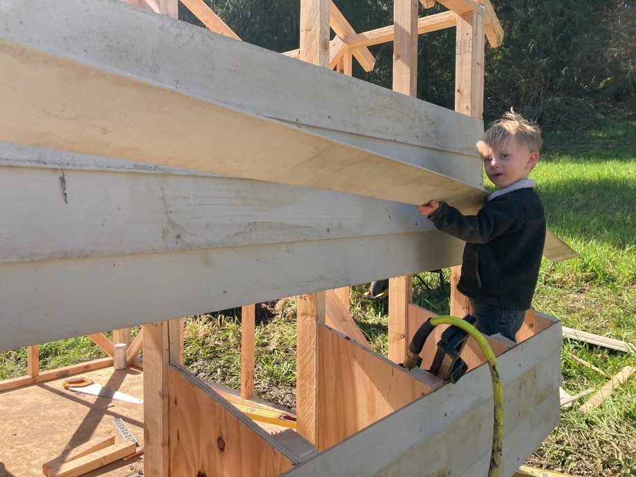 Child in black shirt working on a wooden boat frame outdoors, with beams and grass in the background