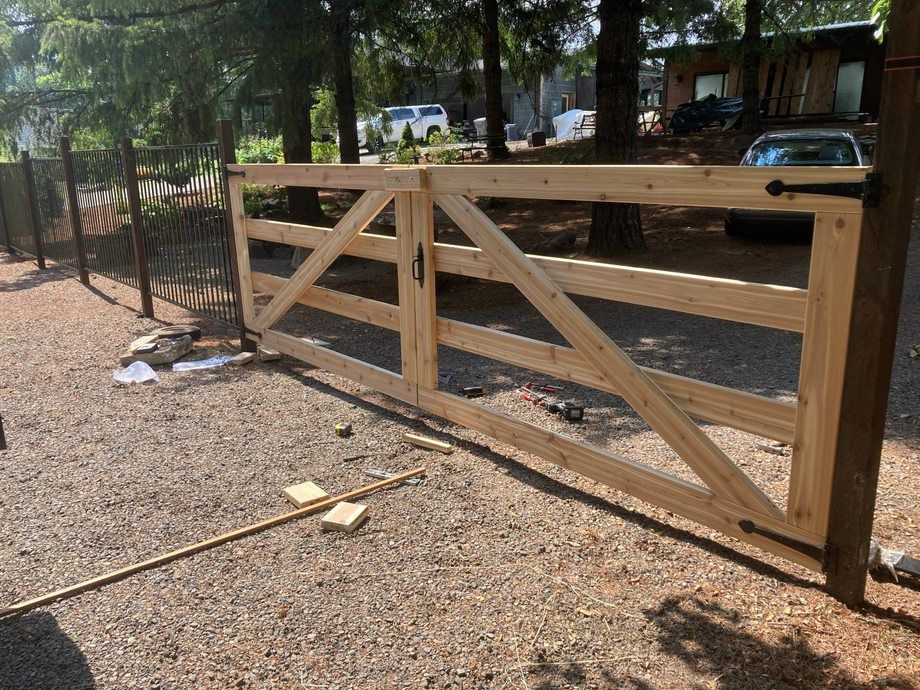 Wooden fence gate with diagonal braces beside a gravel driveway under trees
