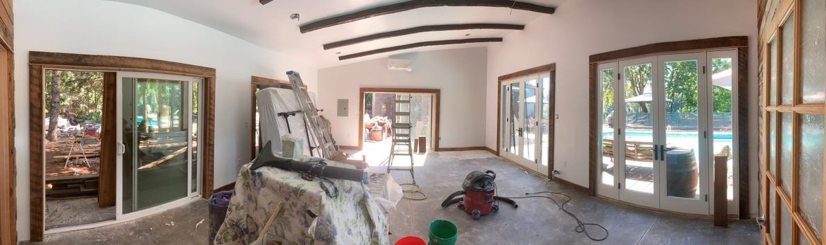 Panoramic view of an unfinished room with exposed beams, stone wall, and large windows/doors letting in daylight