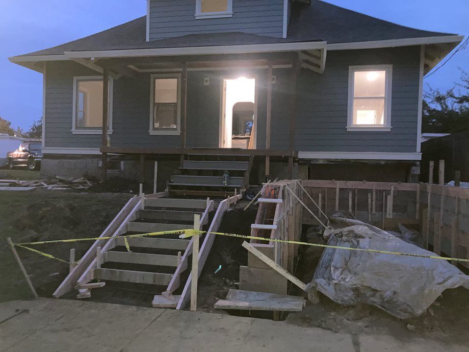 House with front porch under construction, with stairs, scaffolding, and a large rock in the yard.