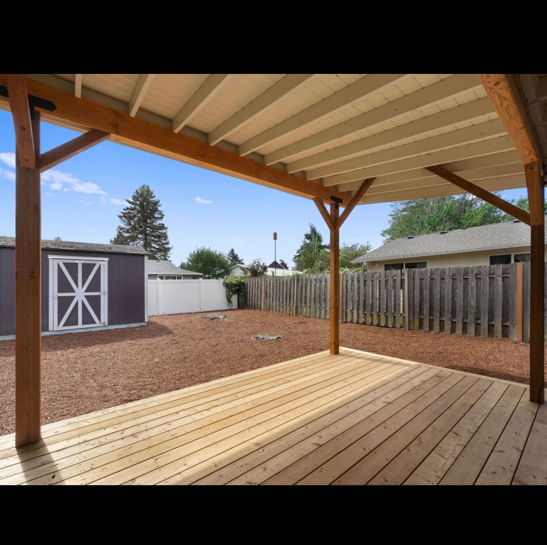 Covered wooden patio overlooking a fenced backyard with a storage shed and gravel yard