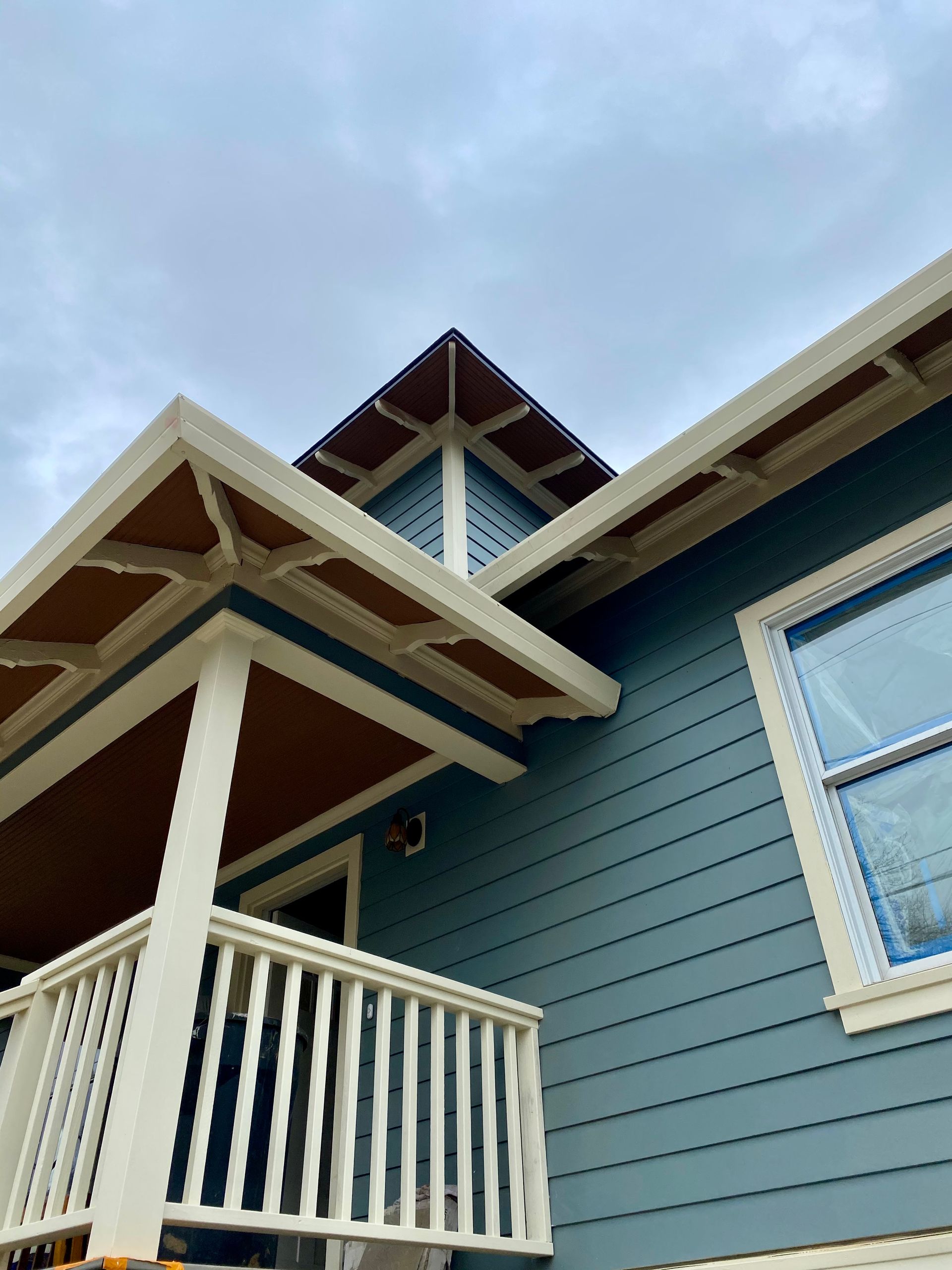 Blue house exterior with white trim, porch railing, and steep gabled roof against a cloudy sky