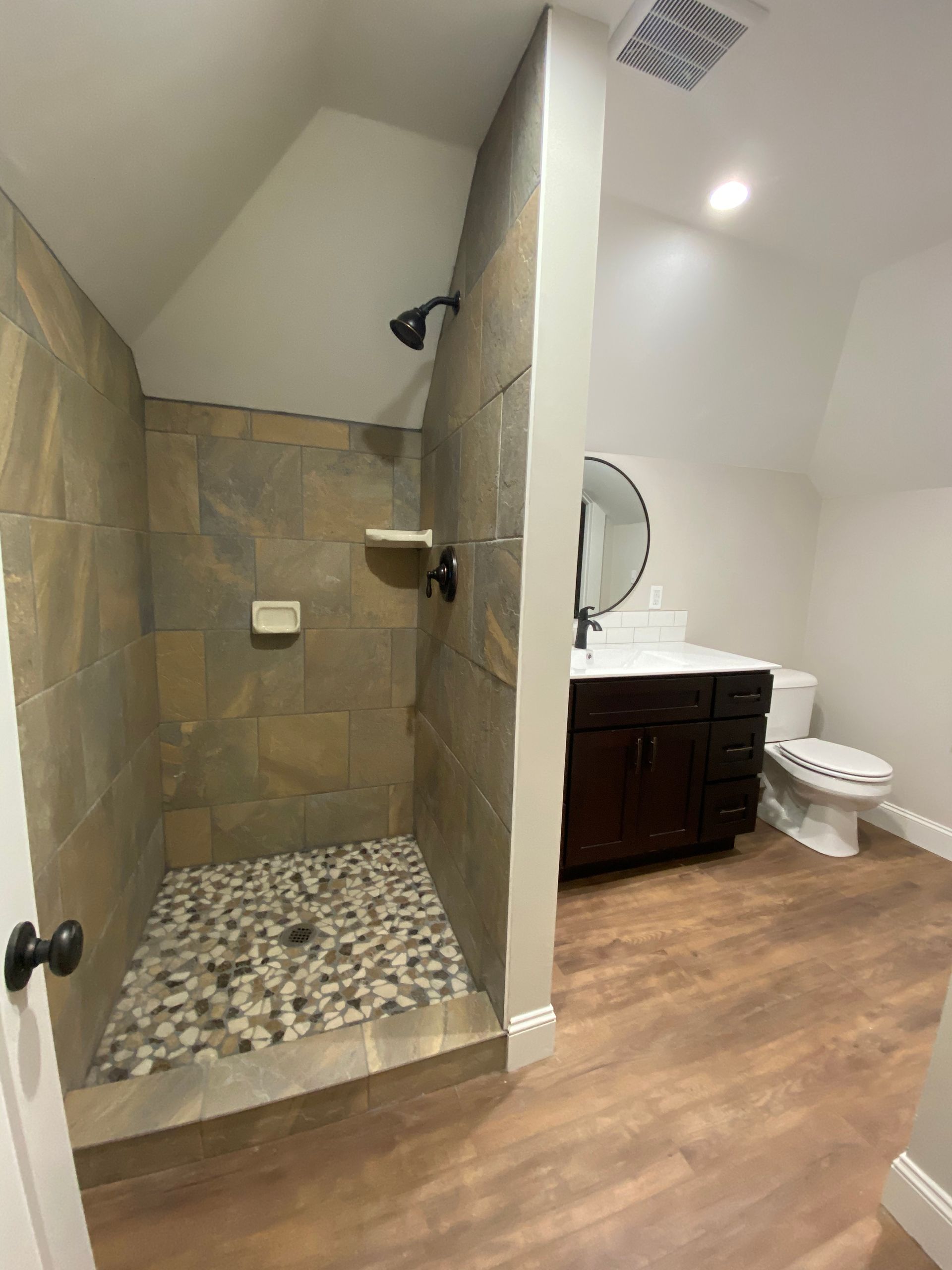 Bathroom with stone-tiled walk-in shower, vanity, and toilet on wood-look flooring.