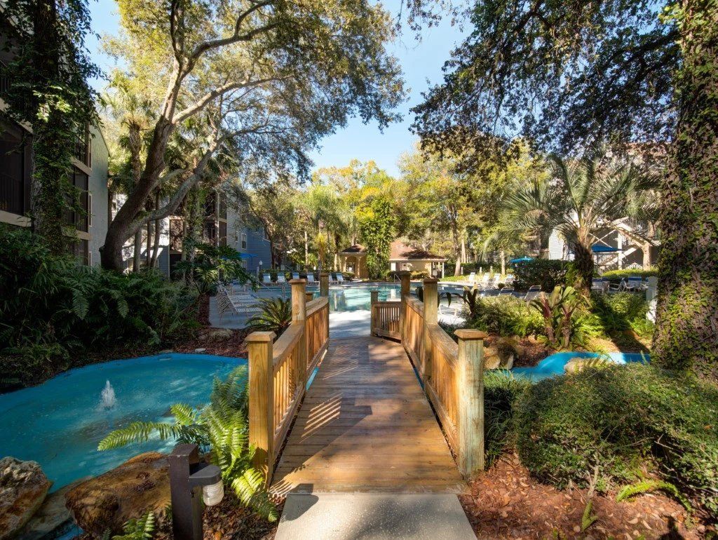 Wooden bridge over a landscaped lagoon with blue water, trees, and lush greenery in a sunny courtyard at Grande Oasis at Carrollwood in Tampa, Florida.