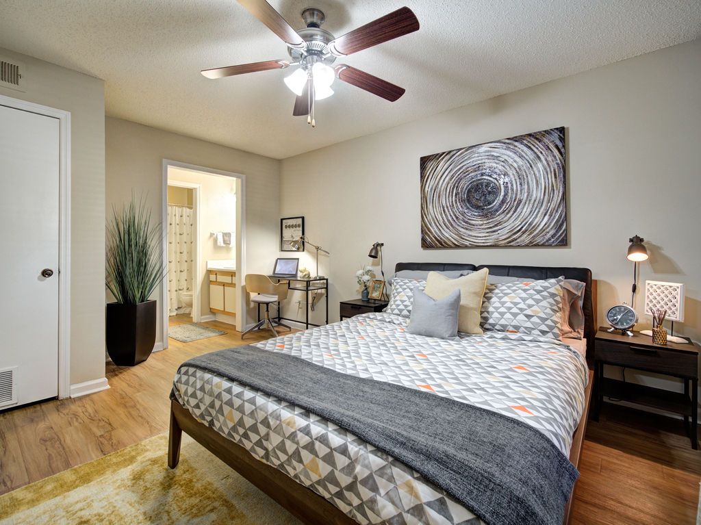 Bright bedroom with a patterned bedspread, ceiling fan, nightstands, desk, and open doorway to a bathroom at Grande Oasis at Carrollwood in Tampa, Florida.