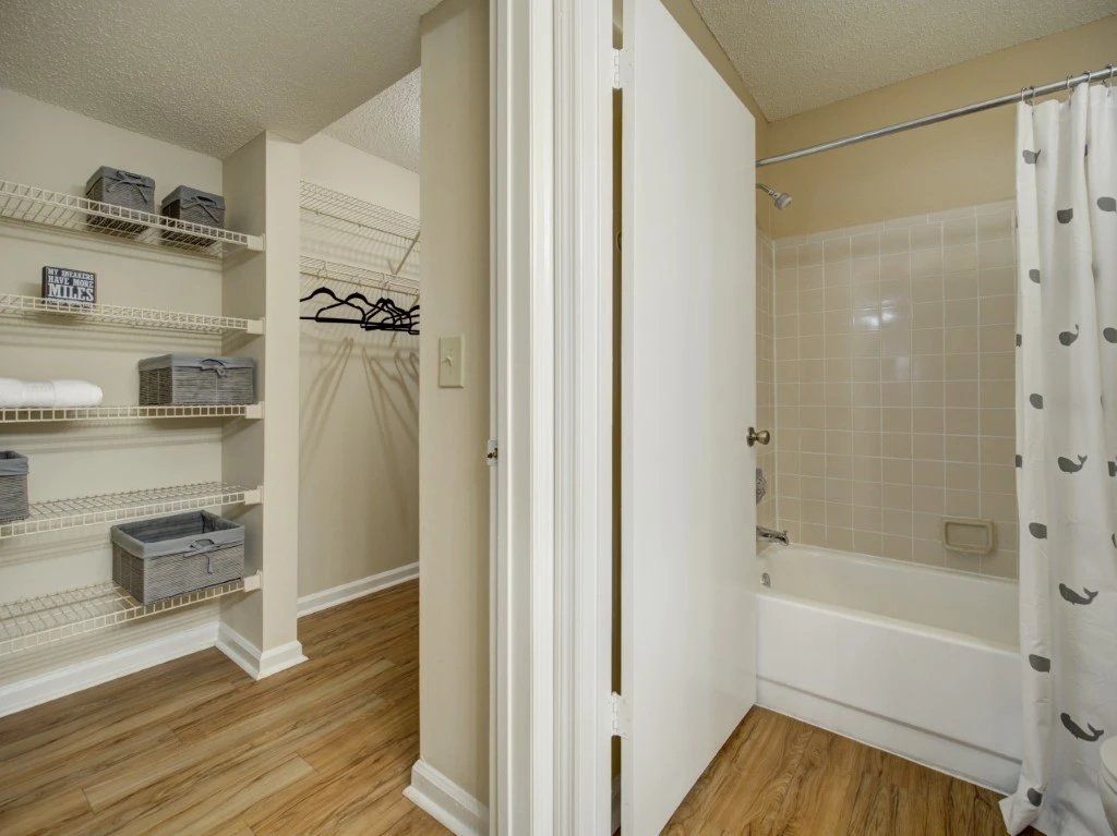 Bright bathroom with white shower curtain, tub, closet shelves, and hardwood floor at Grande Oasis at Carrollwood in Tampa, Florida.