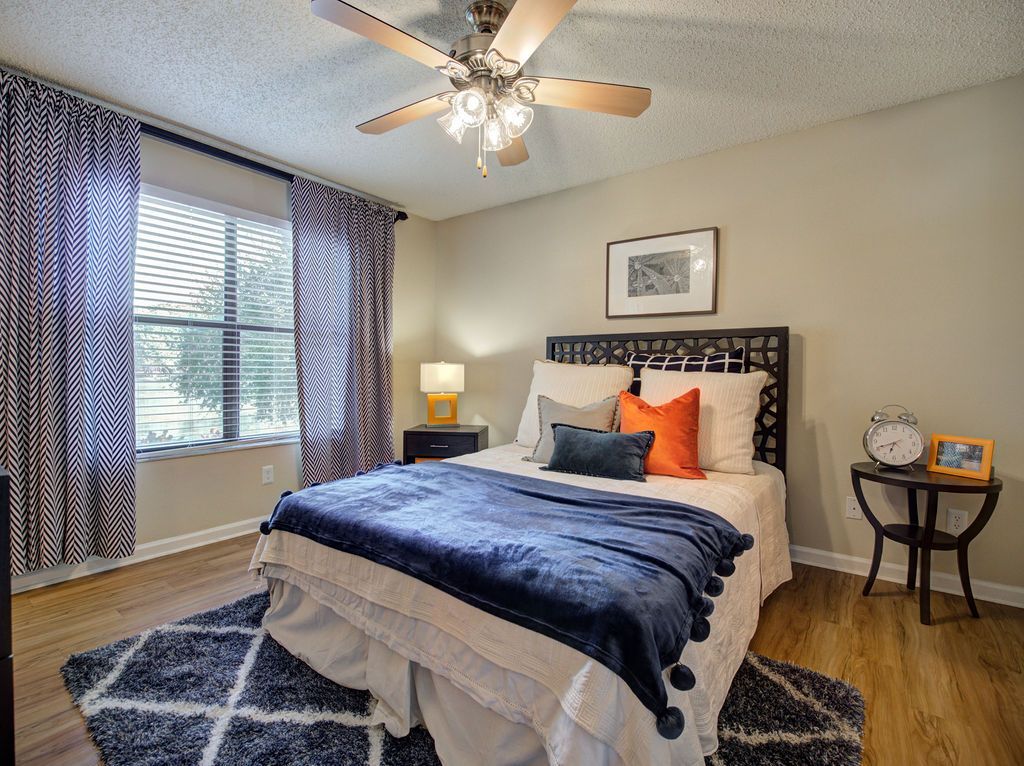 Bright bedroom with a bed, blue blanket, patterned rug, ceiling fan, and large window with curtains at Grande Oasis at Carrollwood in Tampa, Florida.
