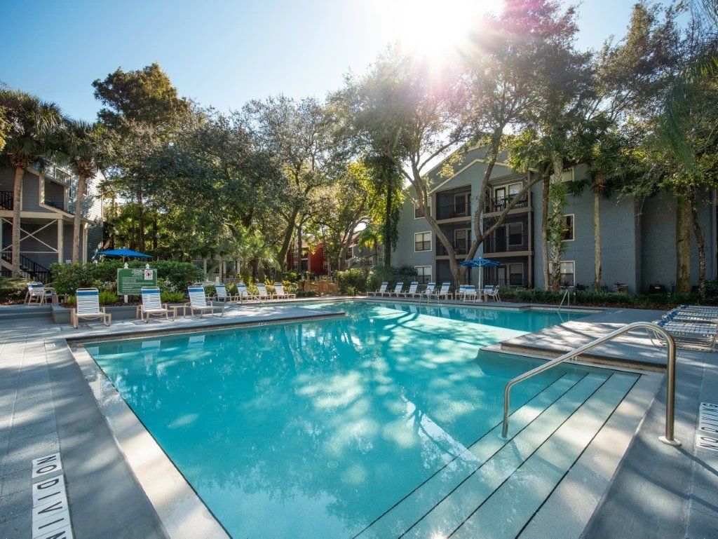 Outdoor resort pool with turquoise water, lounge chairs, and tall trees in bright sunlight at Grande Oasis at Carrollwood in Tampa, Florida.