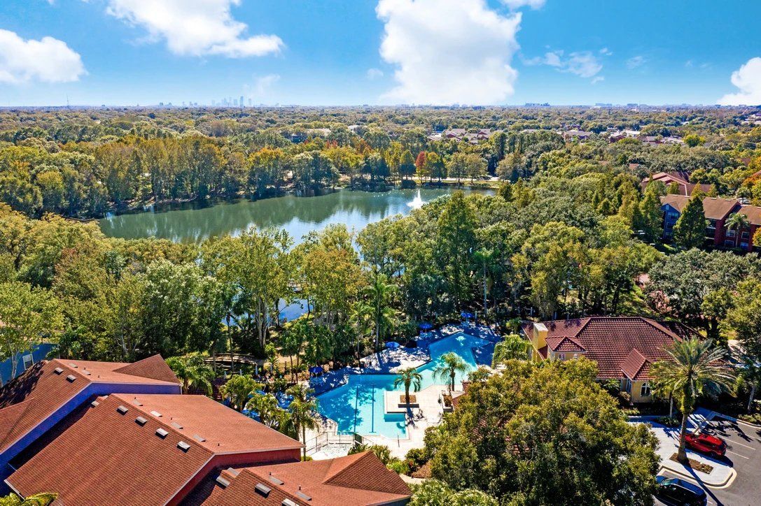 Aerial view of a resort pool, lake, and trees with red-roofed buildings in the foreground at Grande Oasis at Carrollwood in Tampa, Florida.