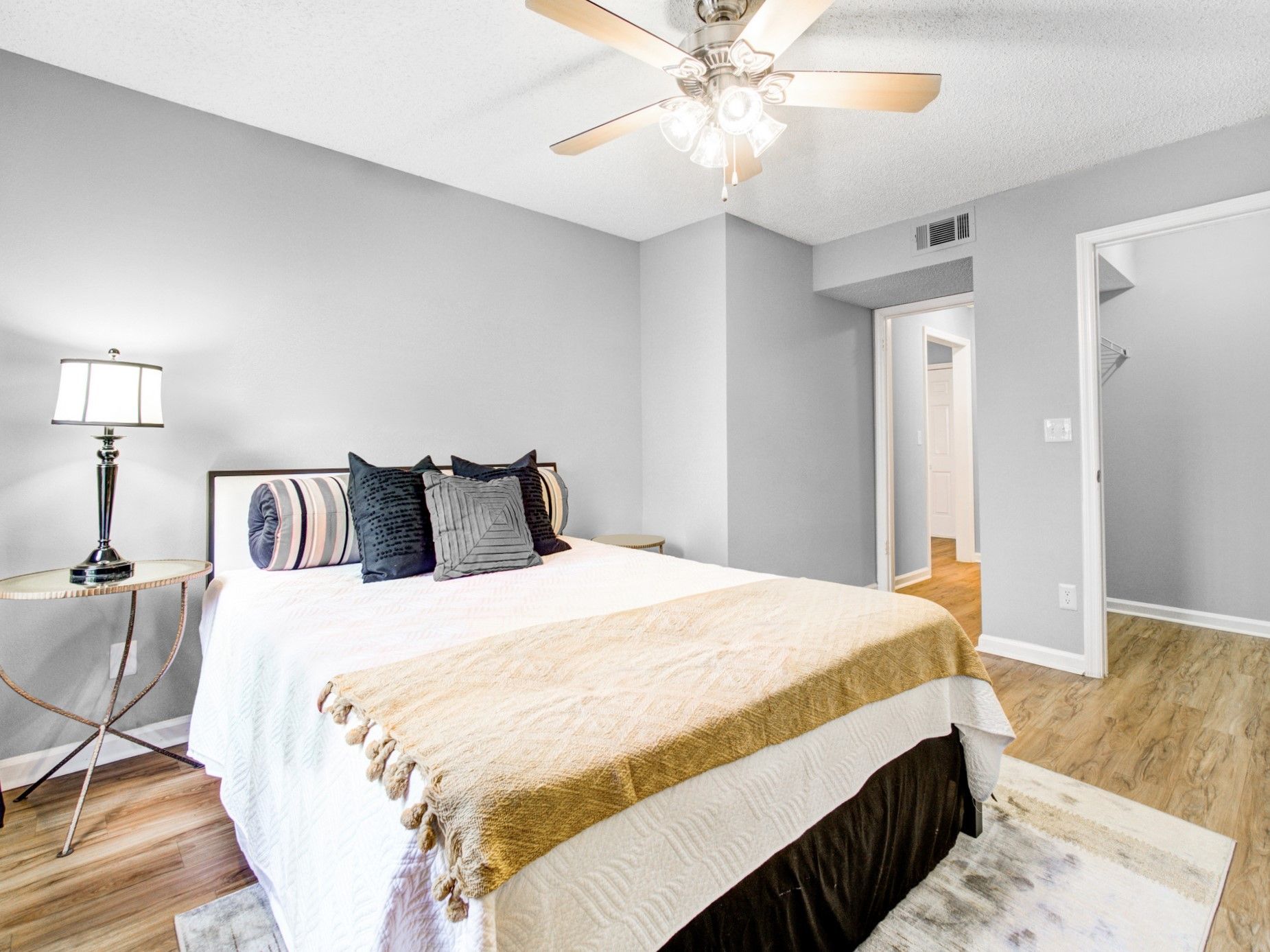 Bright bedroom with a bed, bedside lamp, ceiling fan, and gray walls leading to an open hallway at Grande Oasis at Carrollwood in Tampa, Florida.