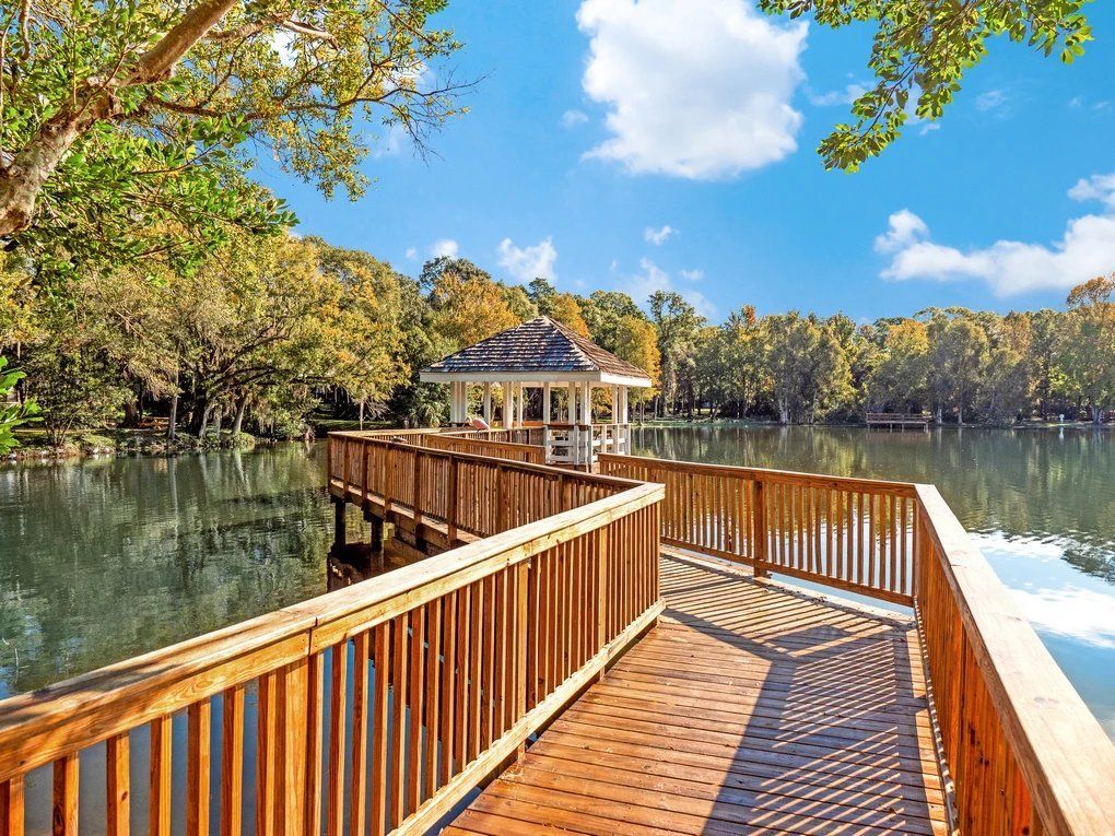 Wooden boardwalk to a lakeside gazebo under a blue sky, surrounded by autumn trees at Grande Oasis at Carrollwood in Tampa, Florida.