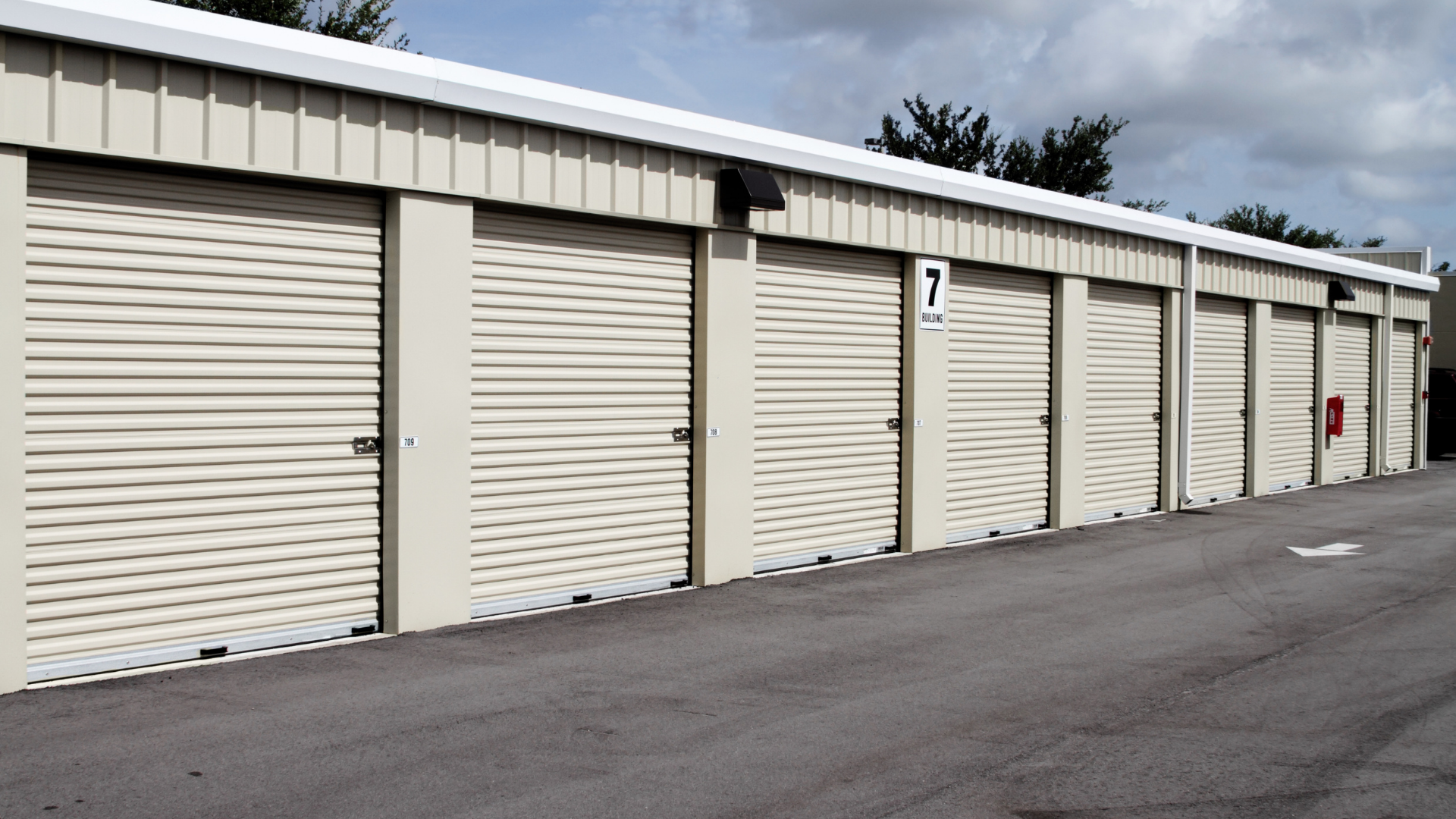 Row of beige storage unit doors along a paved lane, with unit numbers and a red fire extinguisher cabinet.