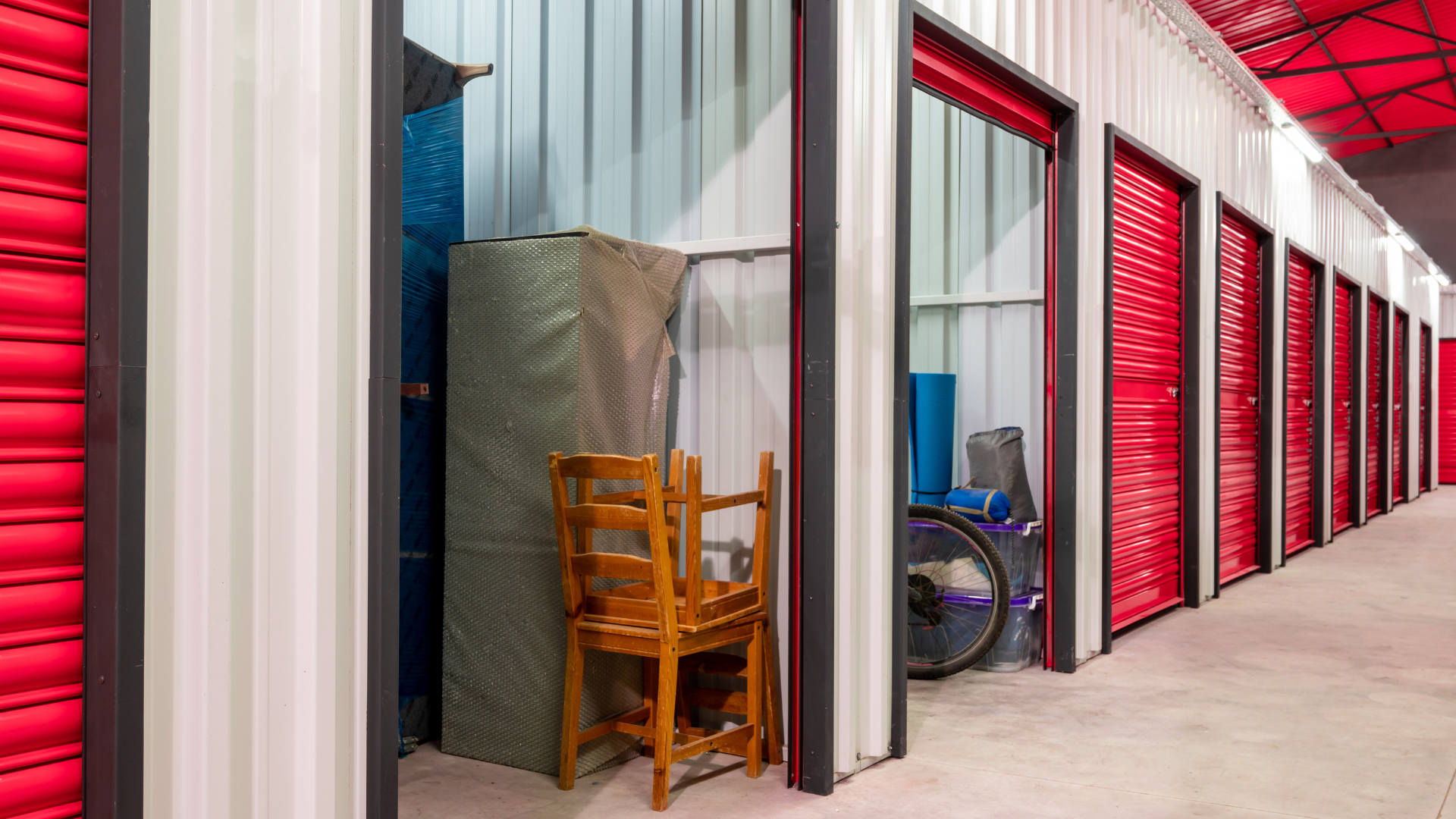 Row of storage units with red doors; one open unit shows stacked furniture and a wheelchair inside.
