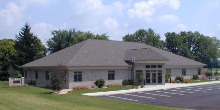 A light brick building with a gray roof and a parking lot on a sunny day.