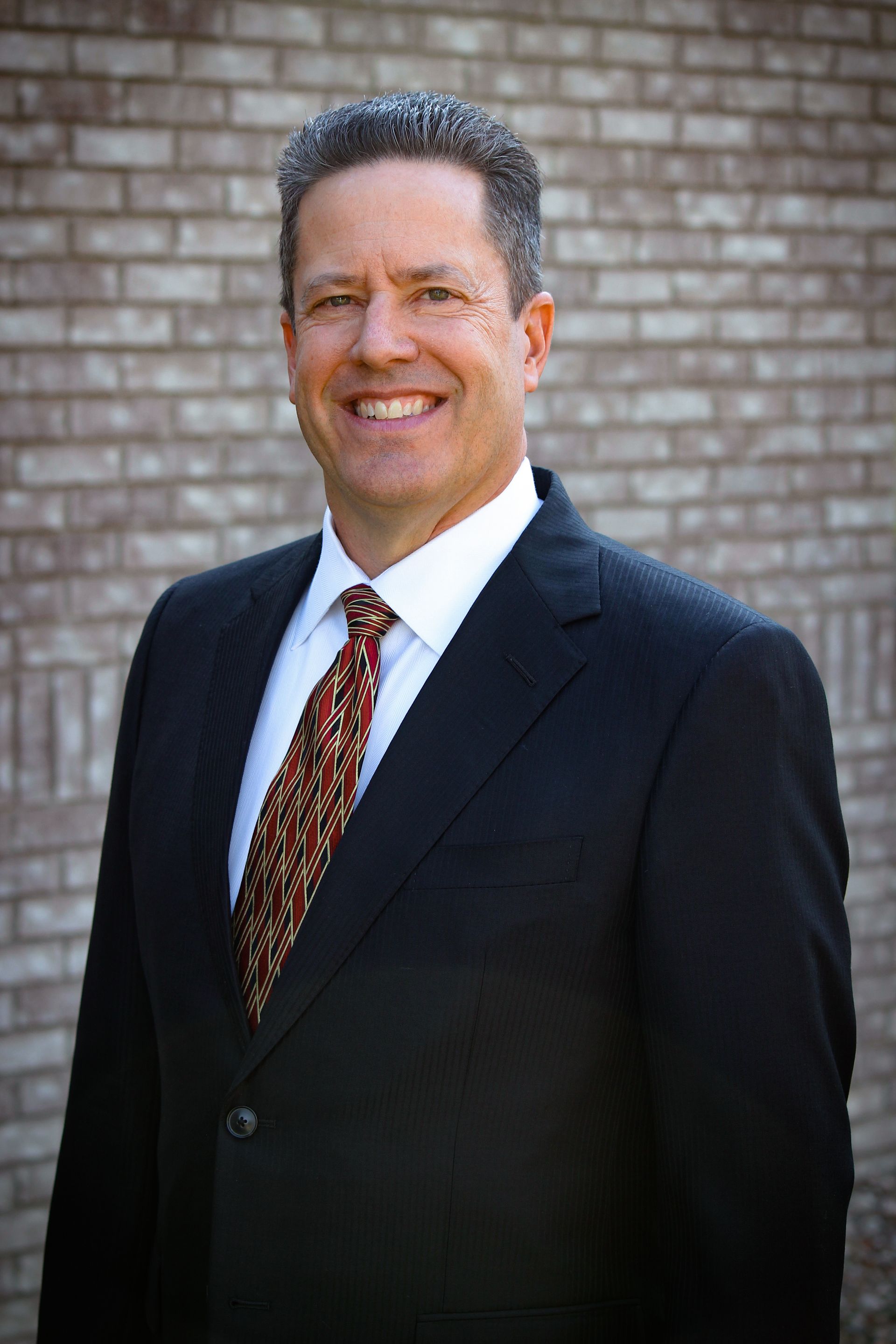 Man in suit smiles in front of a brick wall. He wears a tie with a pattern.