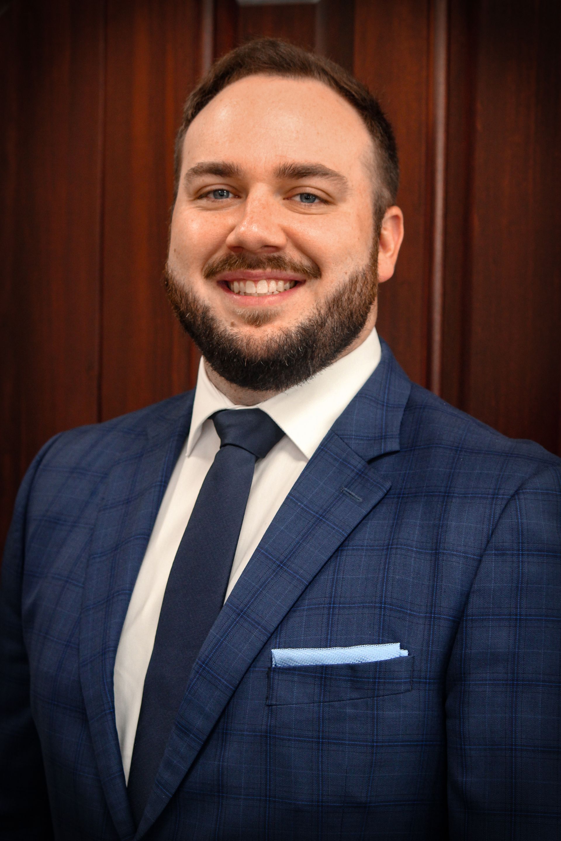 Man in a blue plaid suit, smiling with a dark blue tie and pocket square, in front of wood paneling.