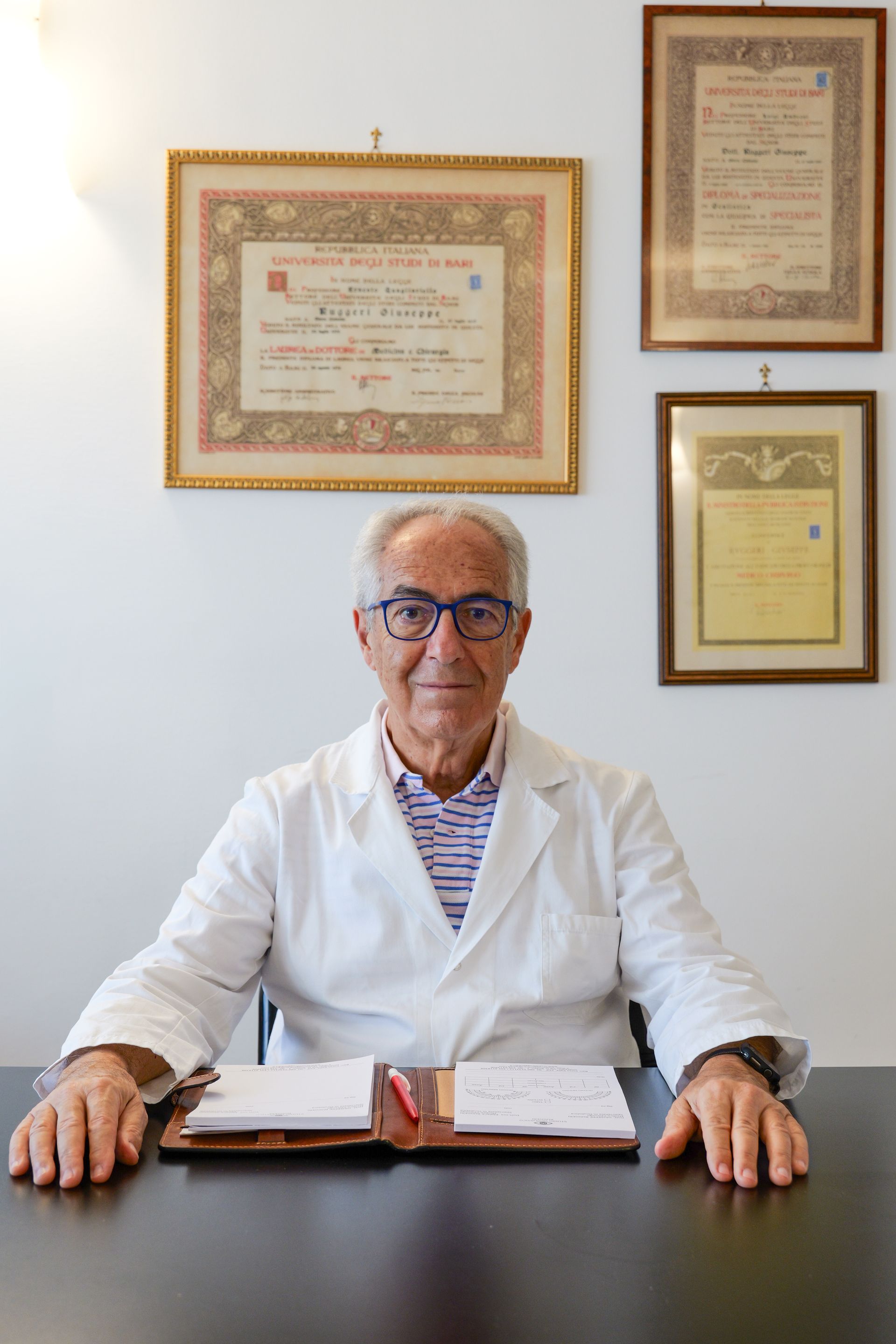 Un medico con occhiali e camice è seduto alla scrivania e guarda la telecamera. Certificati incorniciati sono appesi al muro.