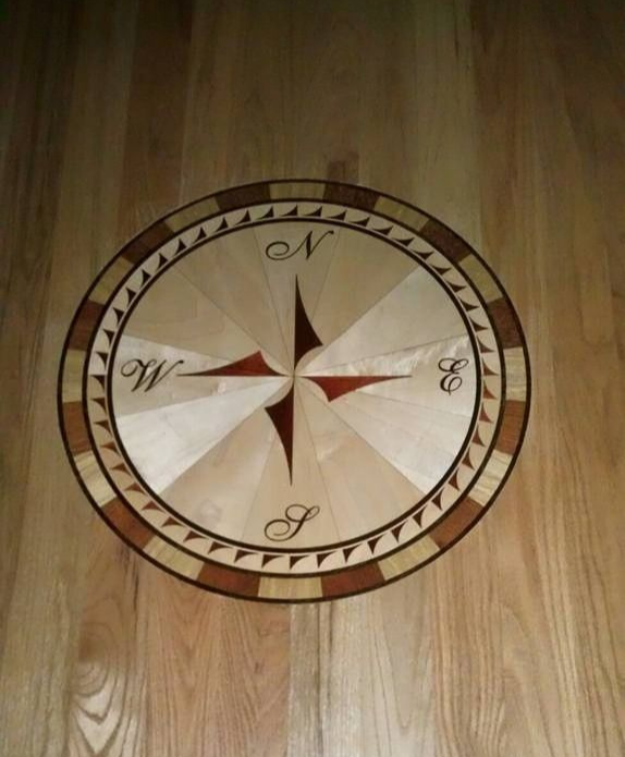 A circular wood compass rose inlay on a light wood floor, featuring dark and light wood segments and directional letters.