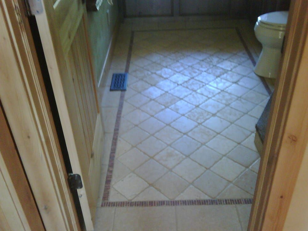 A bathroom floor featuring light-colored diagonal tiles framed by a thin, dark brown border, viewed through a doorway.