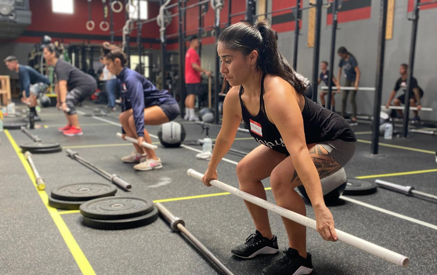 People lifting barbells at a gym. Woman in black tank top focuses on lifting. Others in background.