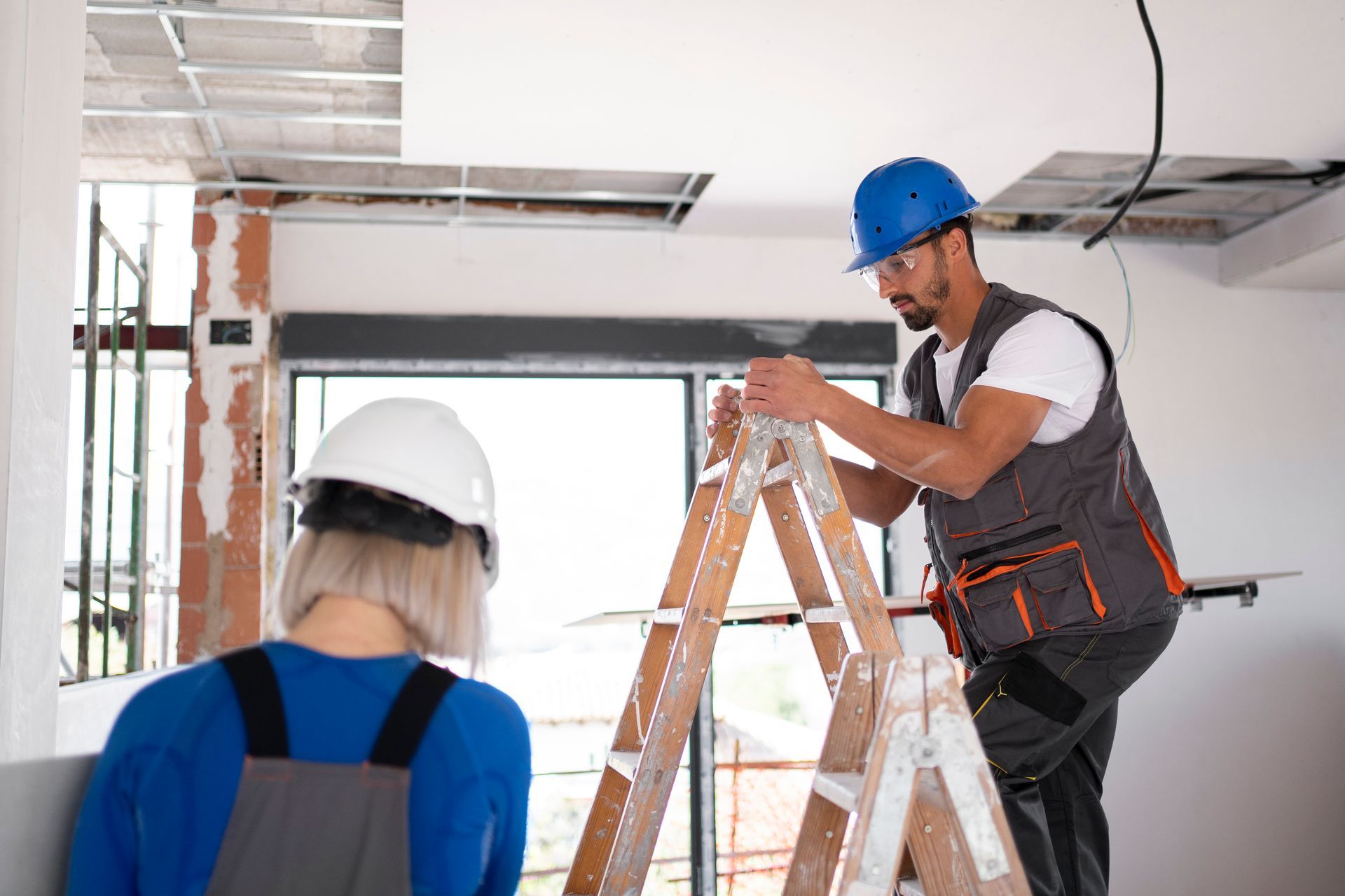 A man and a woman are working on a ladder in a room.