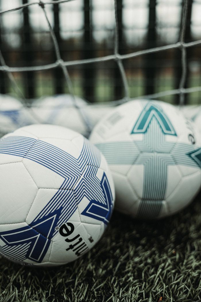Two mitre soccer balls are on the grass in front of a goal