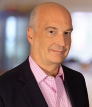 Bald man in black blazer, pink shirt. Smiling, indoor setting.