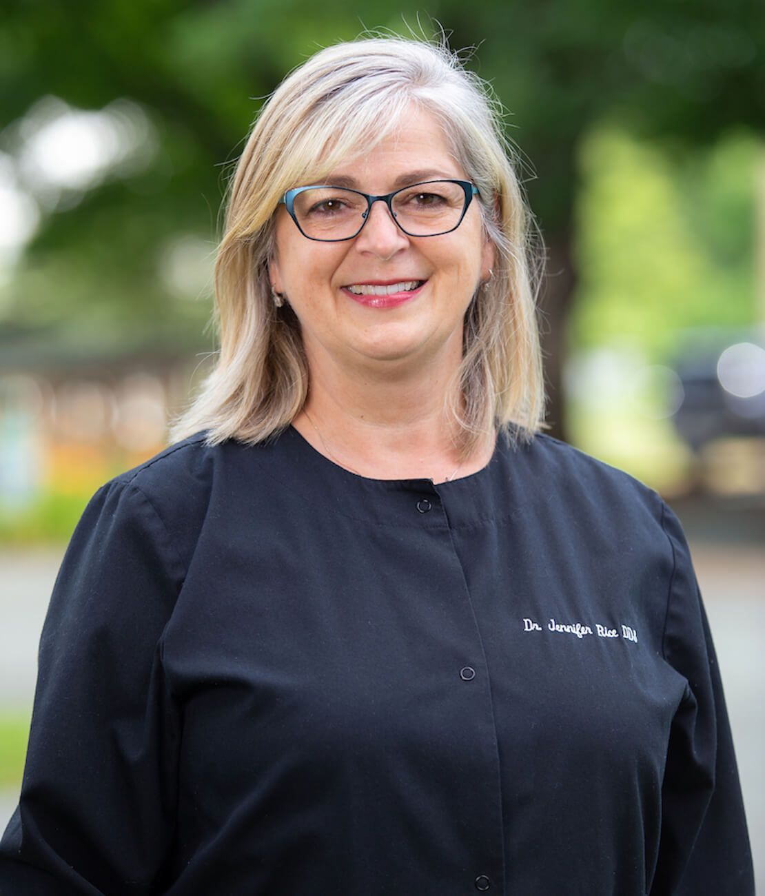 Smiling woman with blonde hair and glasses in black scrubs, outside.