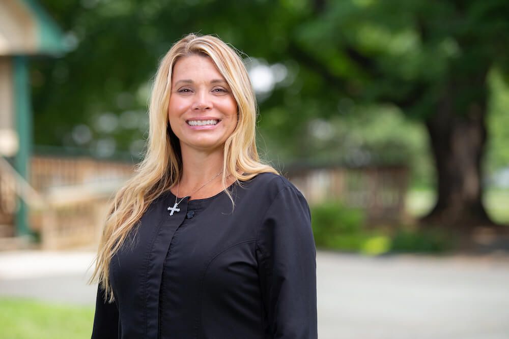 Blonde woman smiling outdoors, wearing a black top with a cross necklace; blurred background.