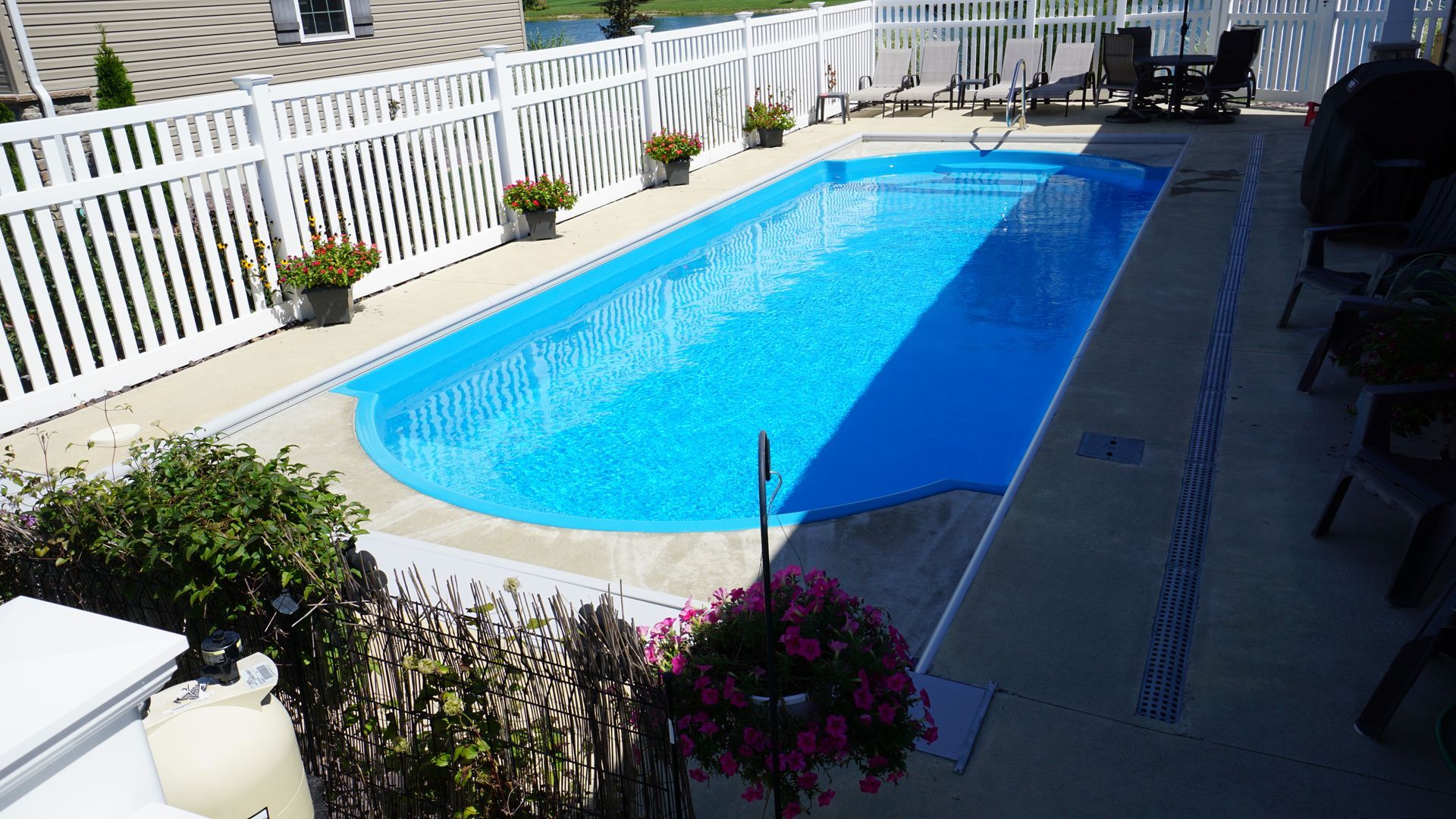 Rectangular pool surrounded by a white fence and patio.  Blue water.  Plants and patio furniture are nearby.