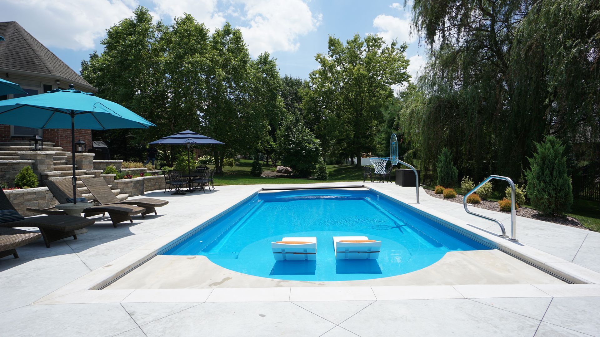 Rectangular blue pool with white coping, lounge chairs, and teal umbrellas on a sunny day.