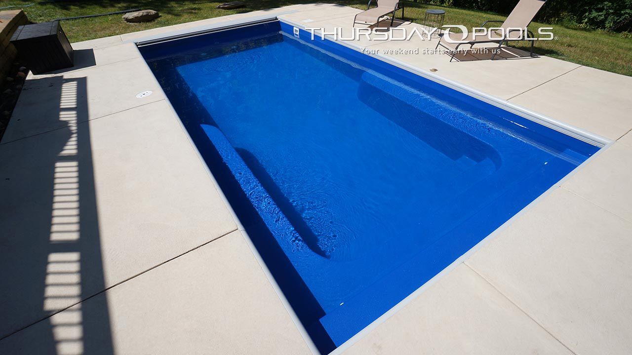 Rectangular blue pool with concrete decking, two lounge chairs in the background, green grass, bright sunlight.