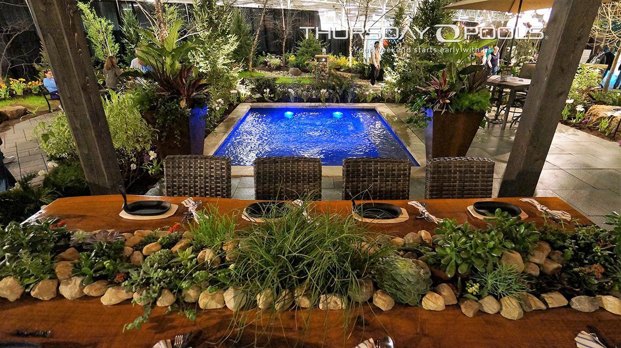 Wooden dining table with stone and plant decor, overlooking a blue lit pool in a garden setting.