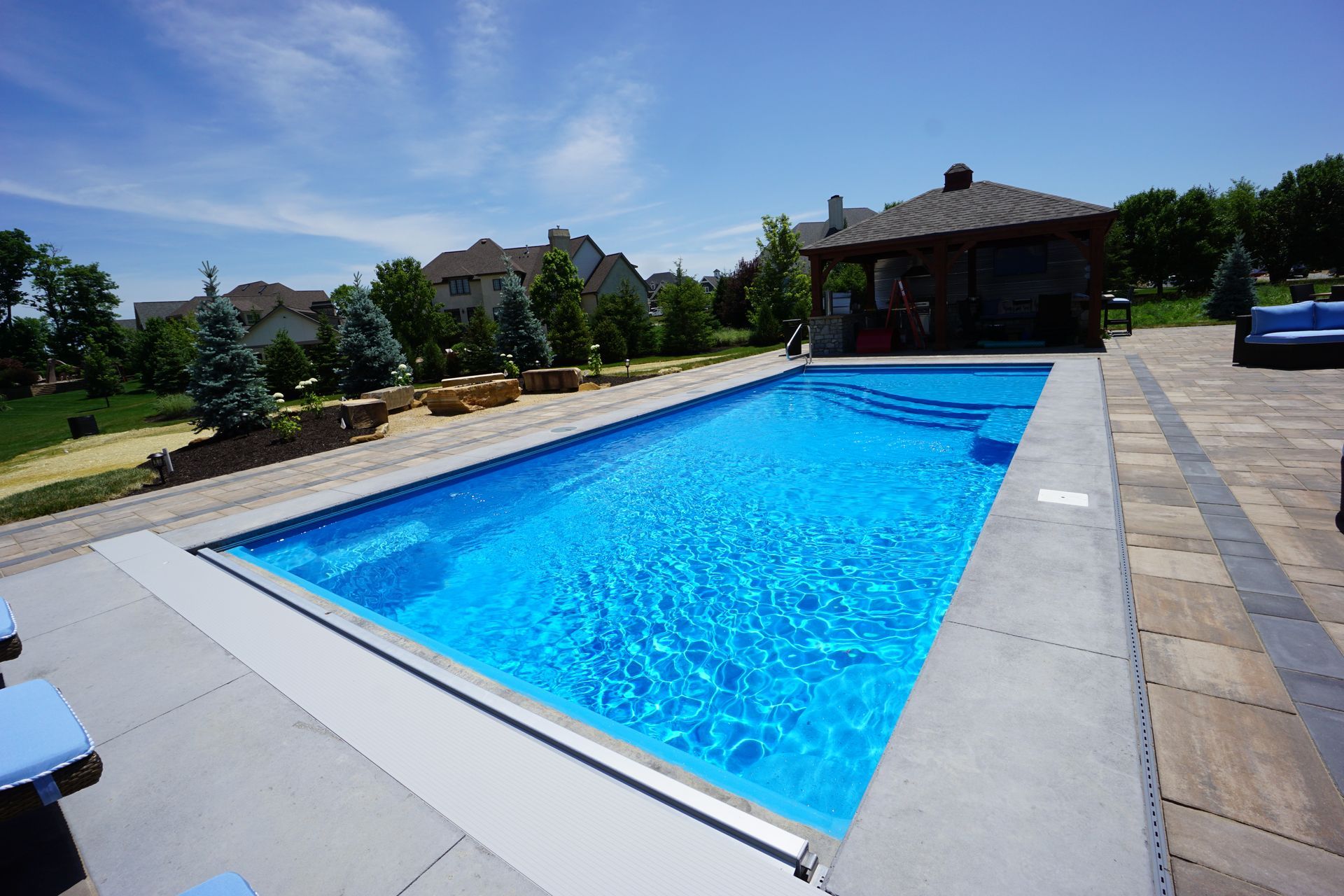 Rectangular blue swimming pool with stairs, patio, gazebo, and houses on a sunny day.
