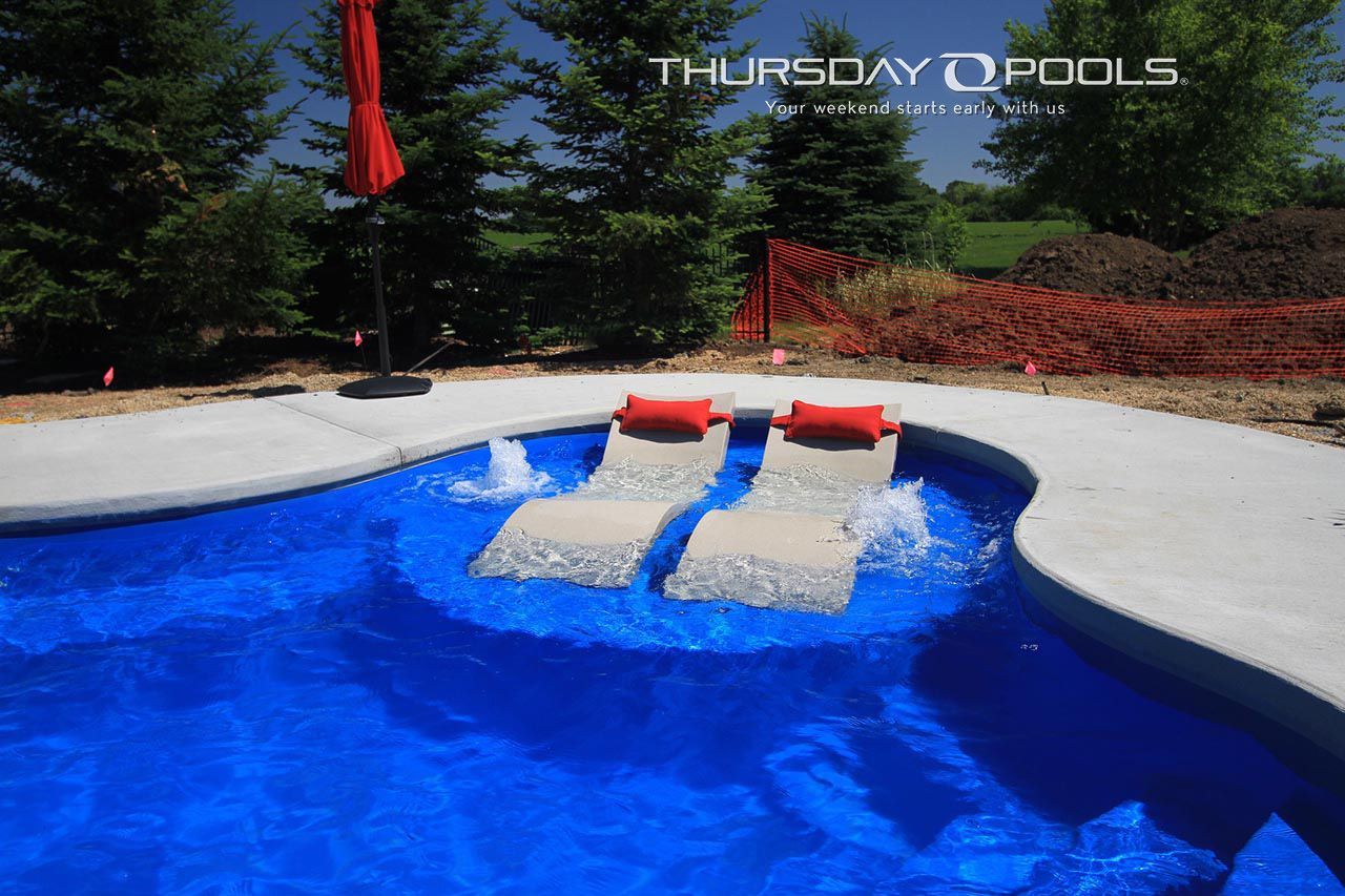 Pool with in-water lounge chairs; blue water; red cushions; concrete deck; sunny outdoor setting.