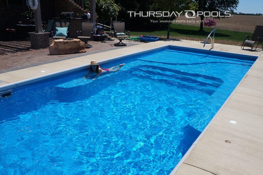 A rectangular blue pool with a person swimming; outdoor setting with patio furniture.
