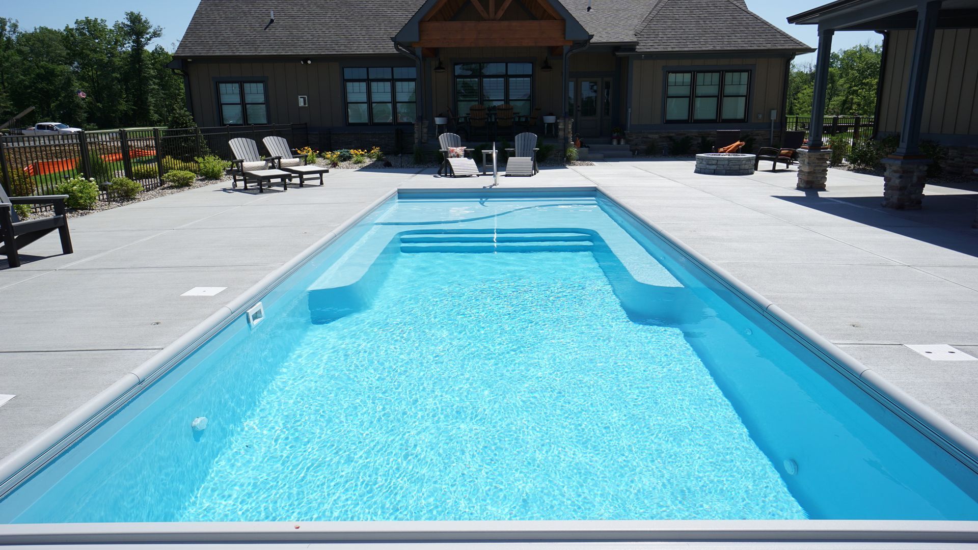 Rectangular turquoise pool surrounded by concrete patio, set against a house with lounge chairs.
