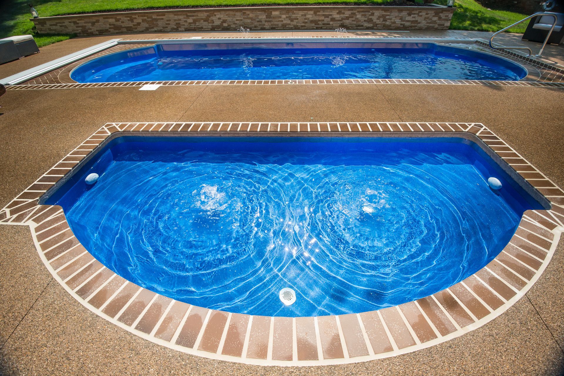 Two blue swimming pools with brick borders surrounded by a concrete patio.