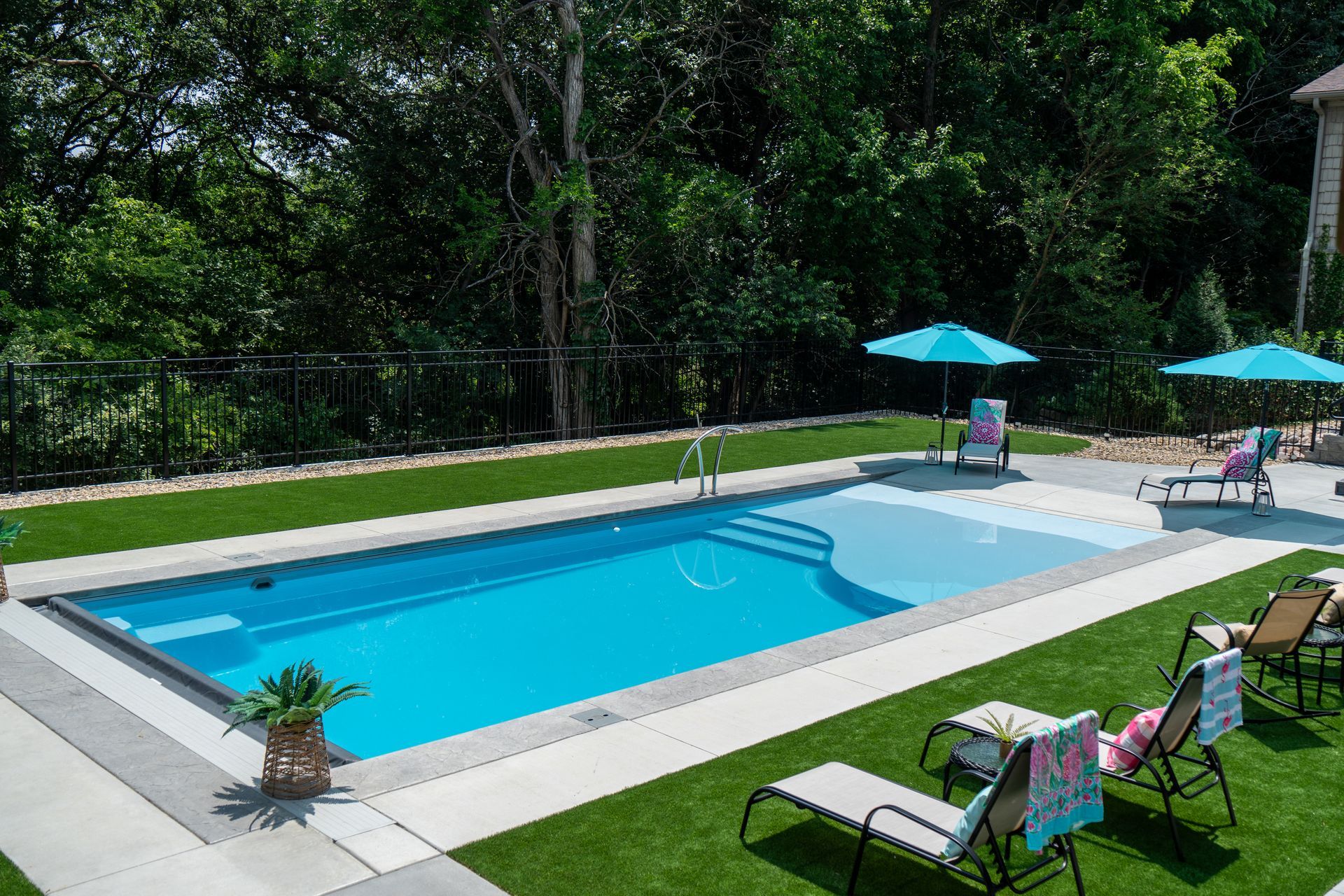 Rectangular in-ground pool surrounded by green lawn and concrete. Teal umbrellas and lounge chairs. Trees in the background.