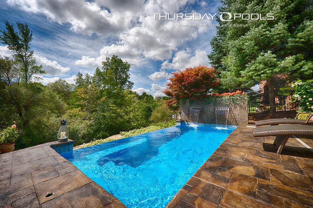 A rectangular swimming pool with turquoise water, a waterfall, and stone patio, surrounded by trees and a cloudy sky.