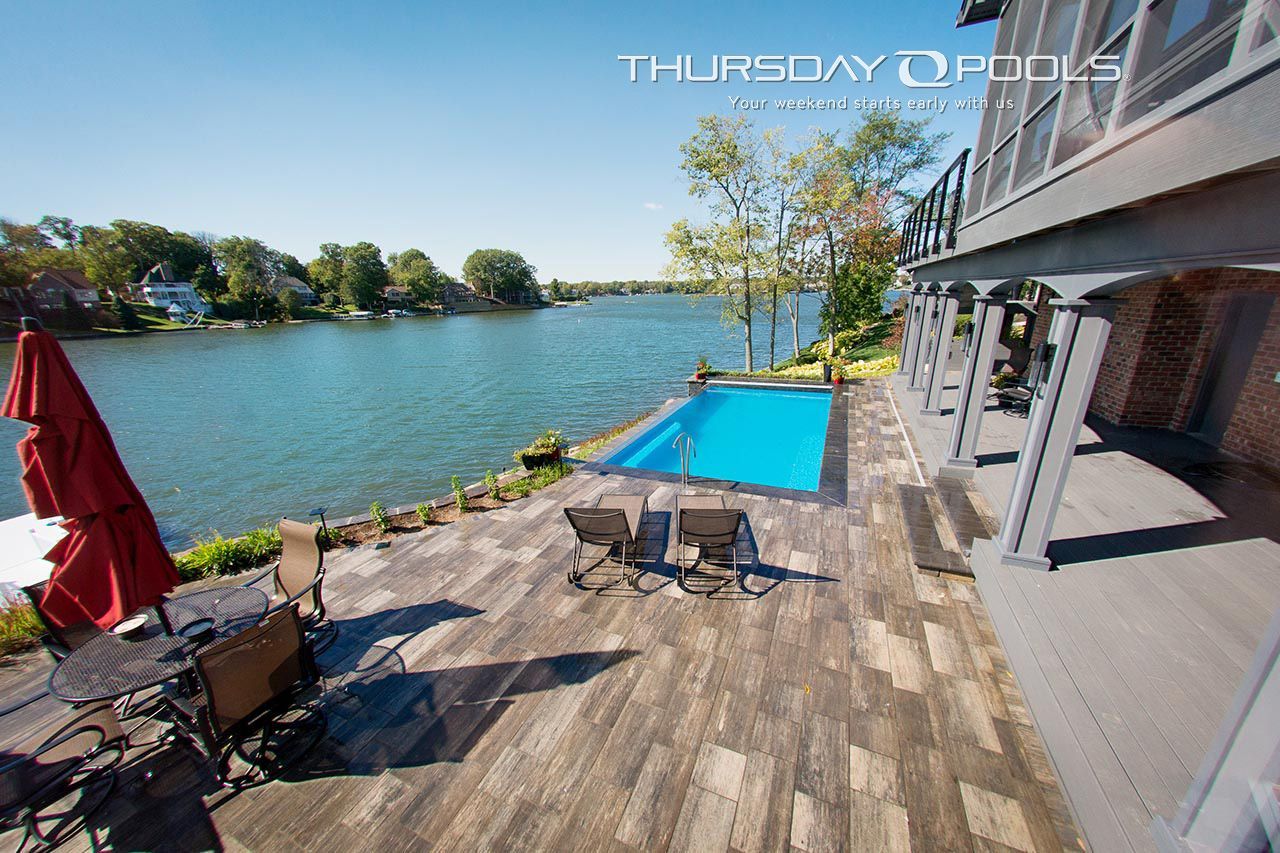 Poolside view overlooking a lake, with a house, patio furniture, and a blue pool.