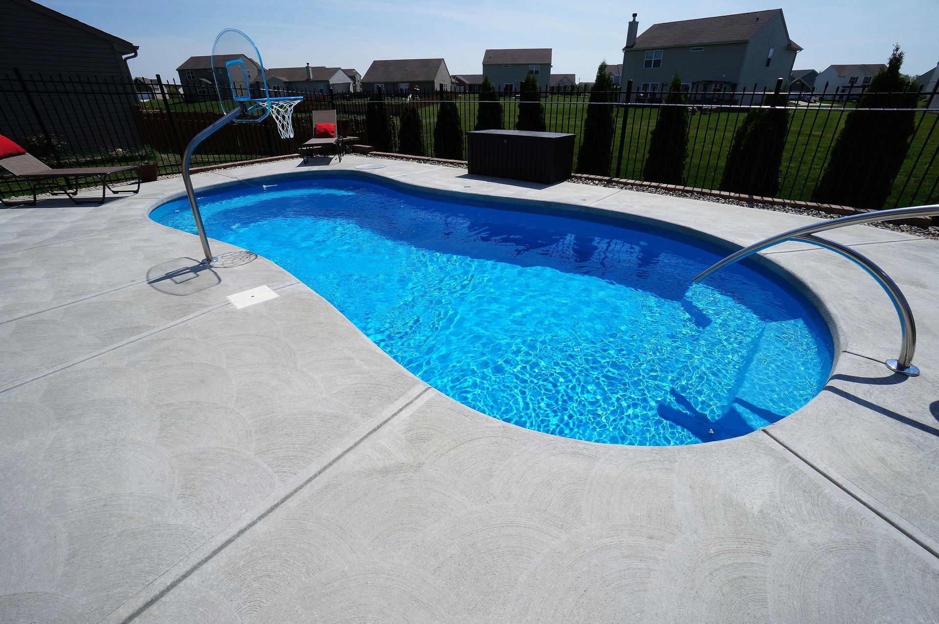 Blue swimming pool with a basketball hoop on a concrete patio. Houses and green hedges in background.