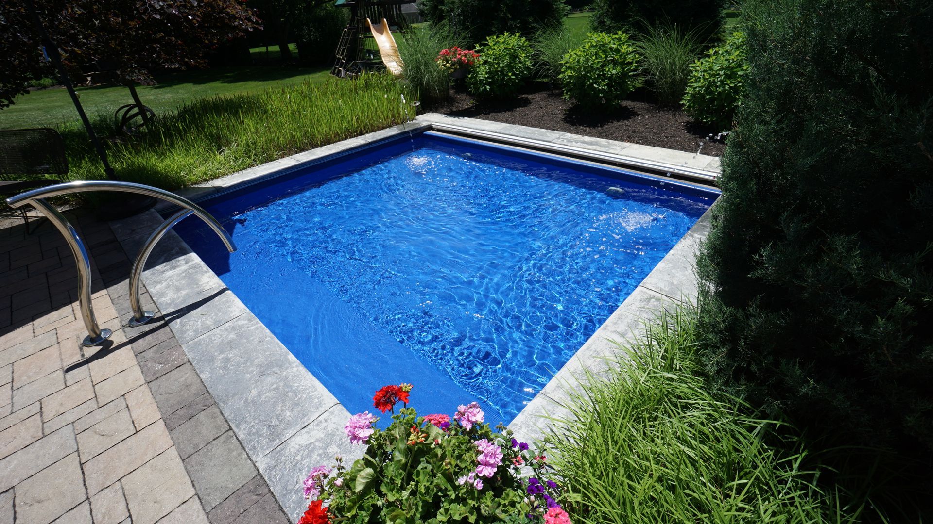 Small, square, blue swimming pool surrounded by stone patio and landscaping.
