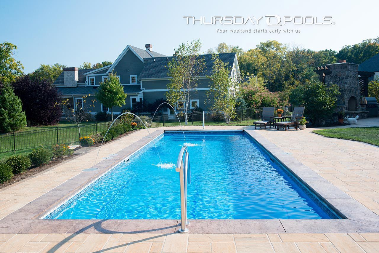 Rectangular swimming pool in a backyard, blue water, stone patio, house in the background, sunny day.